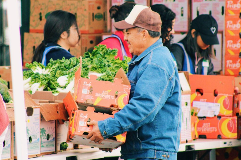 A community member lifts a box of leafy greens from a distribution table as UCHC volunteers prepare and organize produce for attendees. Photo Credit: Photo by Anthony Lujan, Media Outreach Coordinator, Universal Community Health Center