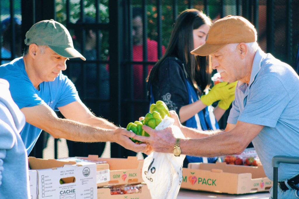 Dr. Chavez, CEO and physician at Universal Community Health Center (UCHC), hands green bell peppers to a community member across a table lined with boxes of fresh produce during a UCHC food pantry event. Photo Credit: Photo by Anthony Lujan, Media Outreach Coordinator, Universal Community Health Center