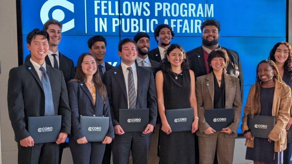 Coro Fellows cohort members holding folders in front of a Fellows Program in Public Affairs display.