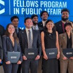 Coro Fellows cohort members holding folders in front of a Fellows Program in Public Affairs display.